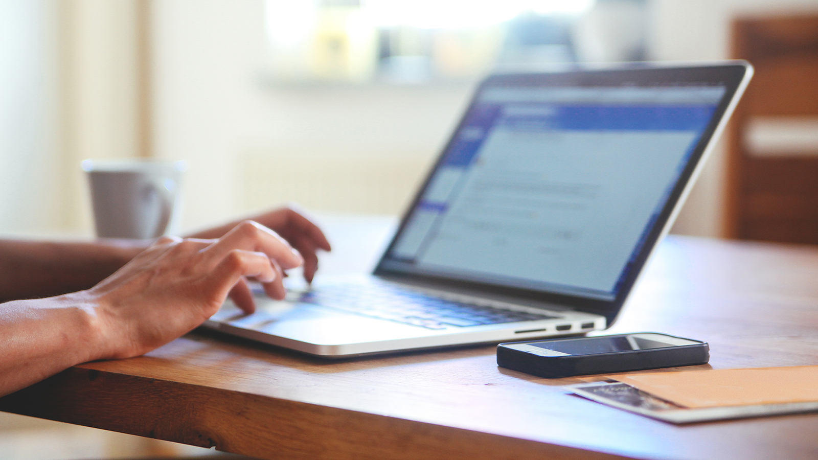 Person at table using a laptop with a smart phone, papers and coffee cup nearby.