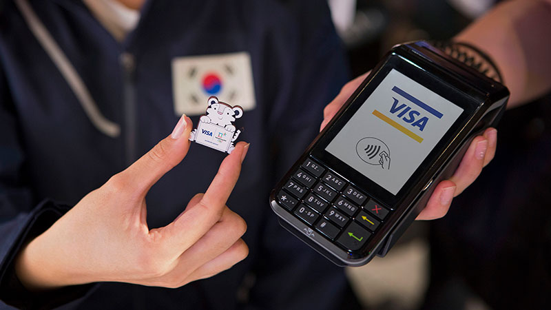 An olympic participant holding a mascot pin and checking out at a store promoting for Visa wearables pin.